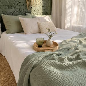 May include: A bed with a white duvet cover and a sage green textured throw blanket. The bed features two sage green pillows, one cream pillow with a small floral pattern, and one cream textured pillow. A round wooden tray holds a green glass.