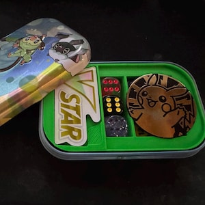 May include: A green and blue tin with a Pokemon Trading Card Game logo and a Pikachu character. The tin is open and reveals a green tray with dice, a VSTAR sticker, and a gold Pikachu pin.