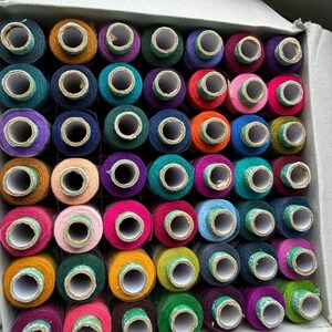 May include: A cardboard box filled with a grid of numerous sewing thread spools in a wide variety of colors including shades of blue, green, pink, purple, orange, yellow, and brown, each featuring a white center core and a small label.