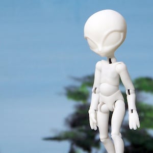 Puede incluir: Una figura articulada de alienígena blanco está de pie sobre una superficie marrón, mirando hacia arriba. Un OVNI plateado y negro borroso flota en el cielo. El fondo presenta un cielo azul y árboles verdes, creando una escena de ciencia ficción.