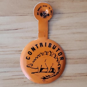 May include: Orange pin with the word "CONTRIBUTOR" and a black illustration of a Stegosaurus. The pin has a small tab at the top with text that reads "Don Howard Assoc NY 10022".