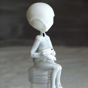 May include: A white articulated doll with a smooth, matte finish, posed in a seated position. The doll has a large head and slender limbs, with its hands clasped. It is sitting on a clear, cylindrical container filled with glitter.