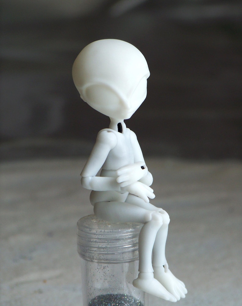 May include: A white articulated doll with a smooth, matte finish, posed in a seated position. The doll has a large head and slender limbs, with its hands clasped. It is sitting on a clear, cylindrical container filled with glitter.