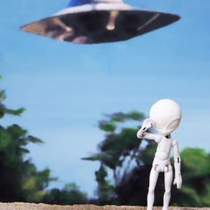 May include: A white articulated alien figure salutes a hovering, metallic-looking UFO against a blue sky backdrop. The scene evokes a sense of wonder and science fiction, with a hint of mystery.