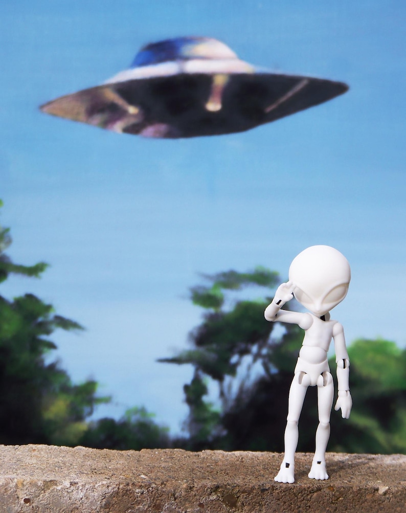 May include: A white articulated alien figure salutes a hovering, metallic-looking UFO against a blue sky backdrop. The scene evokes a sense of wonder and science fiction, with a hint of mystery.