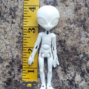 May include: A white articulated alien doll, approximately 4 inches tall, with a large head and slender limbs. The doll has a smooth, matte finish and is posed upright. A yellow measuring tape is positioned next to the doll.