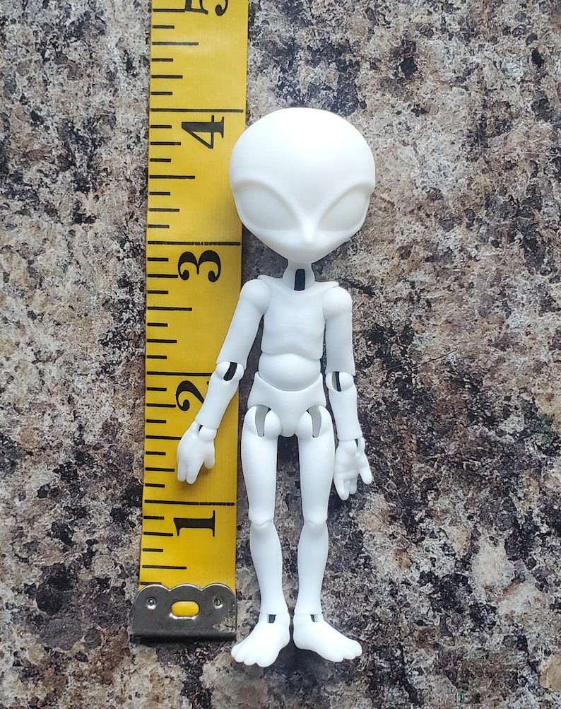 May include: A white articulated alien doll, approximately 4 inches tall, with a large head and slender limbs. The doll has a smooth, matte finish and is posed upright. A yellow measuring tape is positioned next to the doll.