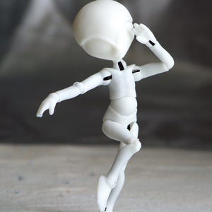 May include: A white articulated doll figure in a dynamic pose, with one arm raised near its head and the other extended. The figure has a rounded head and a slender body, with visible joints. The background is a blurred gradient of gray and white.