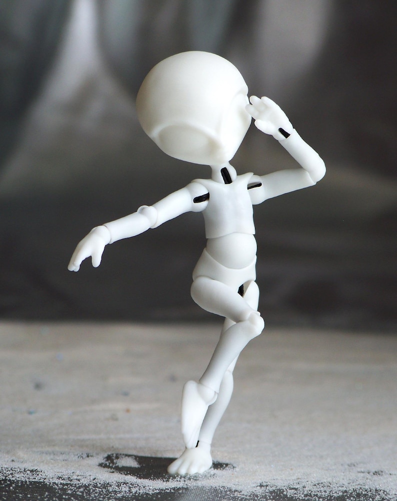 May include: A white articulated doll figure in a dynamic pose, with one arm raised near its head and the other extended. The figure has a rounded head and a slender body, with visible joints. The background is a blurred gradient of gray and white.