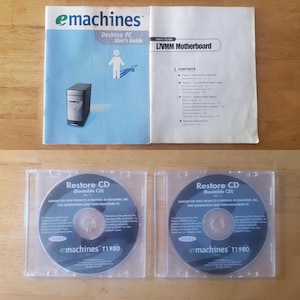 May include: An overhead shot of eMachines computer components. Includes a user's guide, a motherboard guide, and two Restore CDs in clear cases. The user's guide is blue and white, and the CDs have the eMachines logo.
