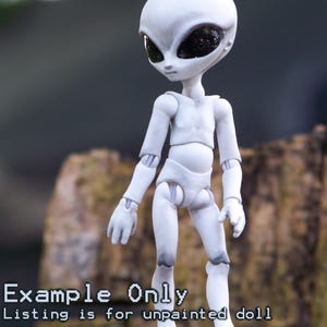 May include: A white articulated alien doll with large black eyes. The doll is standing on a wooden surface. The text "Example Only" and "Listing is for unpainted doll" is displayed on the image.