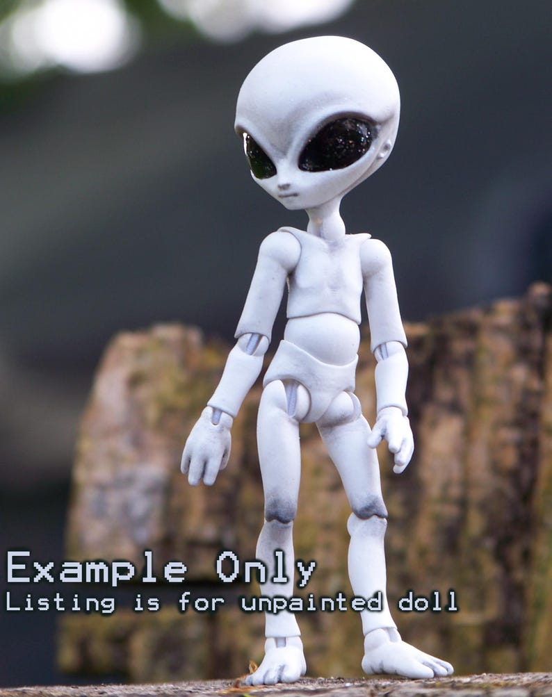 May include: A white articulated alien doll with large black eyes. The doll is standing on a wooden surface. The text "Example Only" and "Listing is for unpainted doll" is displayed on the image.