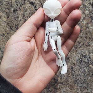 May include: A small, white articulated alien doll with a smooth, matte finish. The doll has a large head with oval eyes and a slender body. The limbs are jointed, allowing for posing. The doll is held in a hand against a mottled brown background.