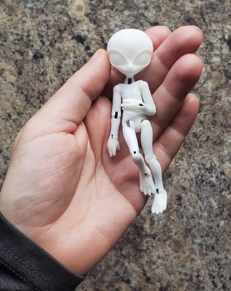 May include: A small, white articulated alien doll with a smooth, matte finish. The doll has a large head with oval eyes and a slender body. The limbs are jointed, allowing for posing. The doll is held in a hand against a mottled brown background.