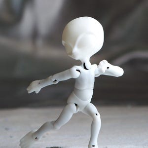 May include: A white articulated doll in a running pose. The doll has a smooth, rounded head and a slender body with visible joints at the elbows, knees, and ankles. The doll's arms are outstretched, and its legs are bent in a running motion.