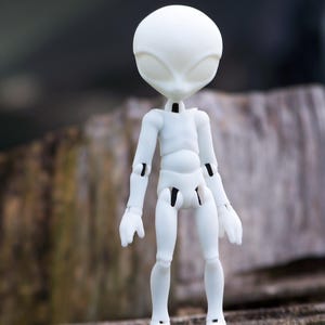 May include: A white articulated alien doll with a large head and oval eyes. The doll has a slender body with jointed arms and legs. The doll is standing on a wooden surface.