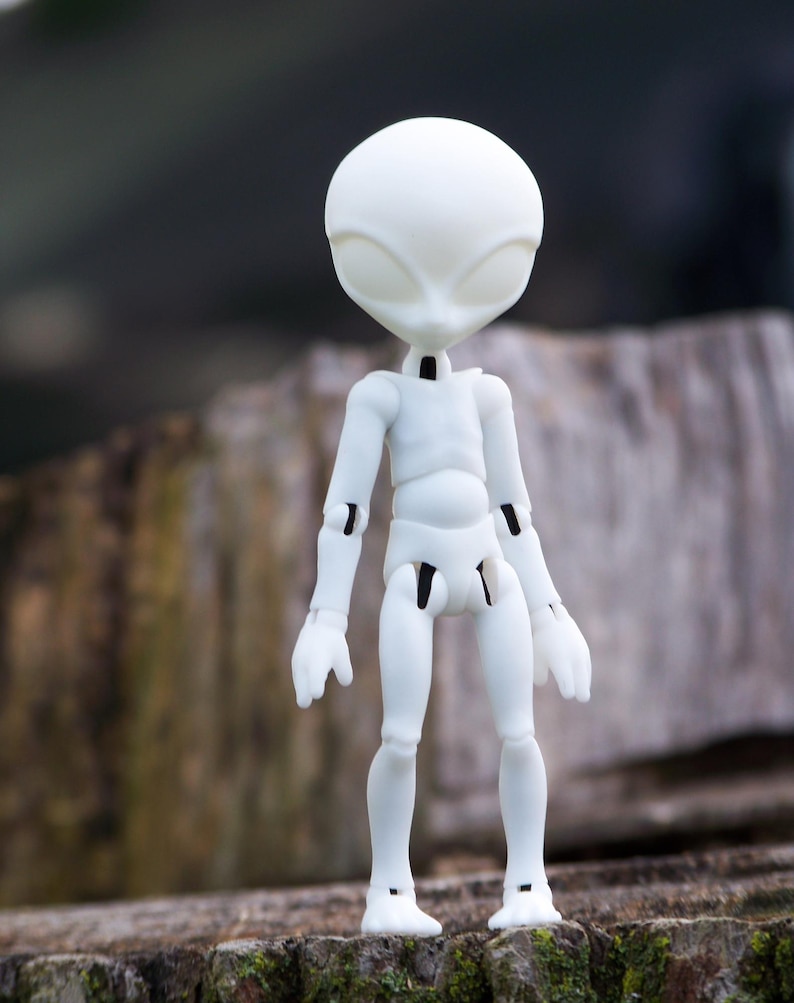 May include: A white articulated alien doll with a large head and oval eyes. The doll has a slender body with jointed arms and legs. The doll is standing on a wooden surface.