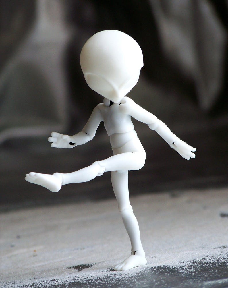 May include: A white articulated doll figure in a dynamic pose, with one leg raised and arms extended. The doll has a smooth, featureless head and body, with visible joints at the elbows, knees, and shoulders. The figure is set against a blurred, neutral background.