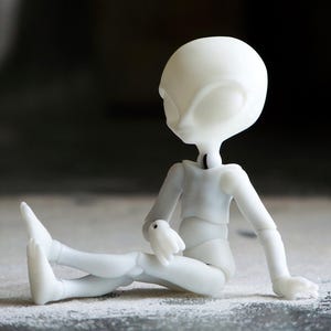 May include: A white, articulated doll in a seated position. The doll has a large head with oval eyes and a slender body. The doll's arms and legs are bent, and it appears to be resting on a textured surface. The doll is made of a smooth, matte material.