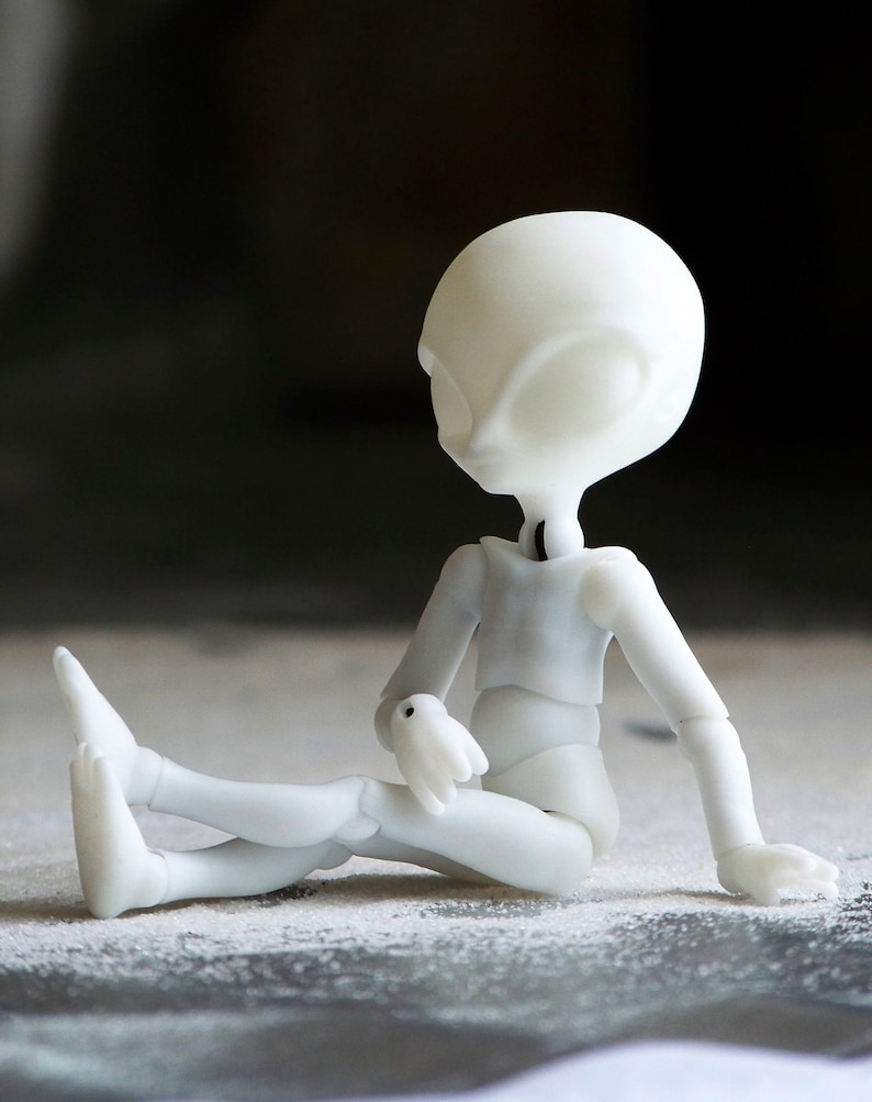 May include: A white, articulated doll in a seated position. The doll has a large head with oval eyes and a slender body. The doll's arms and legs are bent, and it appears to be resting on a textured surface. The doll is made of a smooth, matte material.
