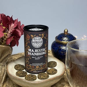 May include: A tin of "Majestic Dianhong" tea coins, with the words "Malty, Velvety, Bright" on the label. The tin is on a small, round, beige dish with several tea coins. A glass of tea is visible.