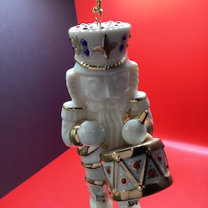 May include: A white porcelain nutcracker ornament with gold and blue accents. The ornament depicts a soldier holding a drum. The drum is light blue with gold trim and red accents. The ornament is suspended by a gold cord.