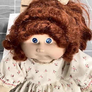 Może przedstawiać: Zabytkowa lalka z rudymi włosami i niebieskimi oczami. Lalka ubrana jest w białą sukienkę we wzory kwiatowe z koronkowym wykończeniem. Kremowa kokarda zdobi włosy lalki. Twarz lalki ma lekko smutny wyraz.