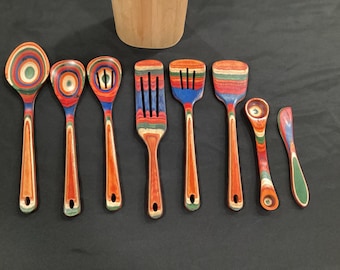 May include: A collection of eight wooden kitchen utensils with colorful, striped patterns. The set includes a spoon, slotted spoons, a spatula, a spoon, and a spreader. The handles are a natural wood color, and the utensils have a hole for hanging.