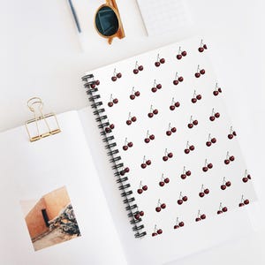 May include: A white spiral bound notebook with a repeating pattern of red cherries on the cover.