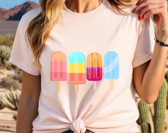 May include: A light pink t-shirt featuring four colorful popsicle illustrations. The popsicles are in shades of orange, blue, yellow, and pink, each with a wooden stick. The shirt is knotted at the waist and worn with blue jeans.
