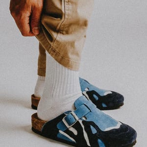 Puede incluir: Zuecos de ante azul y negro con diseño blanco y cierre de hebilla. Los zuecos tienen una plantilla de corcho. Llevados con calcetines blancos y pantalones color caqui.