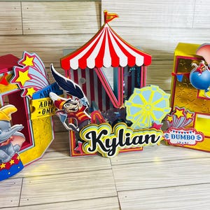 Può includere: Decorazione a tema circo colorata con Dumbo l'elefante, un tendone da circo e una ruota panoramica. Il design include il nome "Kylian" e la parola "Dumbo". La tavolozza dei colori comprende rosso, giallo, blu e oro.