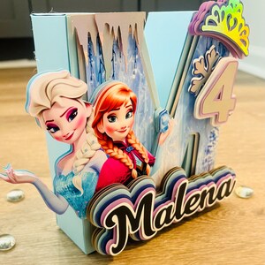 May include: A light blue, multi-layered birthday decoration featuring the characters Elsa and Anna from Frozen. The decoration includes the number 4, a crown, and the name "Malena" in a layered design. The overall theme is a birthday celebration.