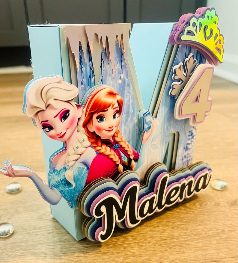 May include: A light blue, multi-layered birthday decoration featuring the characters Elsa and Anna from Frozen. The decoration includes the number 4, a crown, and the name "Malena" in a layered design. The overall theme is a birthday celebration.
