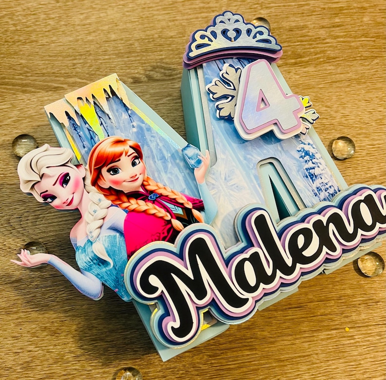May include: A layered birthday cake topper featuring the characters Elsa and Anna from Frozen. The topper includes the number 4, a crown, a snowflake, and the name "Malena" in a layered design. The color scheme is primarily blue, white, and purple.