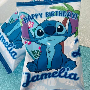 May include: Blue and white party favor bags featuring a cartoon character with large ears and big eyes. The bags have the text "Happy Birthday!" and the name "Jamelia" in a script font. The background has a tropical theme with palm trees and flowers.