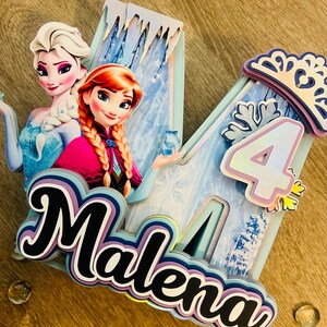 May include: A layered cake topper featuring the characters Elsa and Anna from the movie Frozen. The design includes the name "Malena" and the number "4", with a snowflake and crown. The color scheme includes blue, purple, and white.