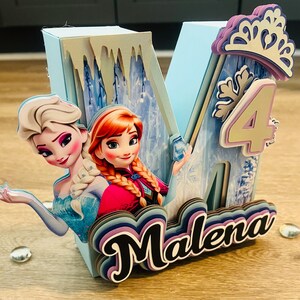 May include: A light blue, layered letter "M" with the name "Malena" in a cursive font. The letter features images of Elsa and Anna from Frozen, a number "4", and a crown. The design is for a birthday.