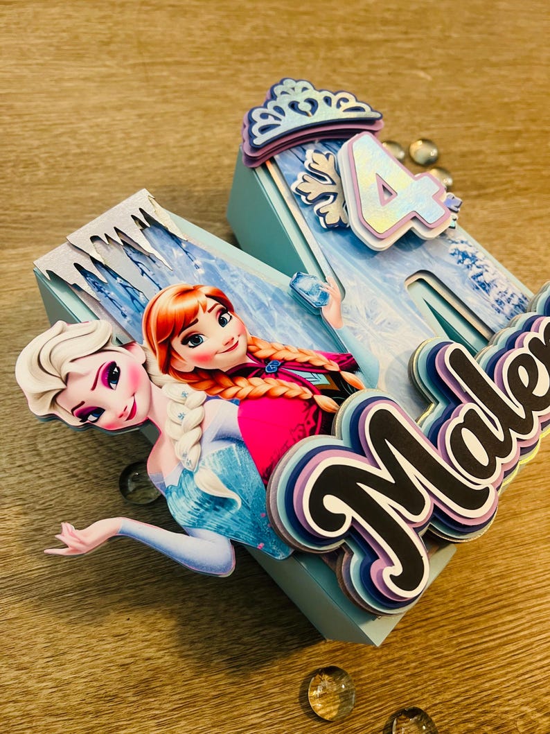 May include: A light blue, layered birthday decoration featuring the characters Anna and Elsa from Frozen. The number 4 is displayed, along with the name "Malena" in a layered design. The decoration is set on a wooden surface.
