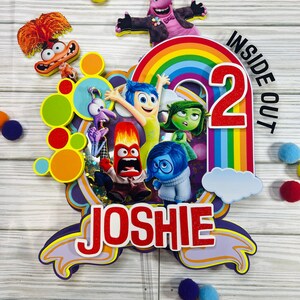 May include: A vibrant cake topper with characters from "Inside Out". It features a rainbow, the number 2, and the name "Joshie" in red. The design includes character cutouts and colourful pom-poms, perfect for a birthday celebration.