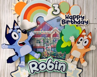 May include: A colorful cake topper featuring cartoon characters, a rainbow, and the words "Happy Birthday" and "Robin". The design includes a house, sun, and the number "3". The characters are in shades of blue and orange.