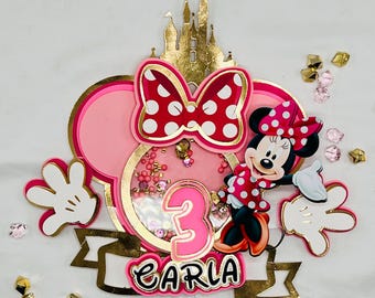 Könnte beinhalten: Minnie-Maus-Geburtstagskuchenaufsatz. Das Design zeigt ein rosa-goldenes Farbschema, eine Minnie-Maus-Figur, eine Schleife, die Zahl 3 und den Namen "Carla". Der Aufsatz ist mit goldenen Akzenten und Konfetti verziert.