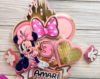 Könnte beinhalten: Ein rosa-goldener Tortendekor mit einer Zeichentrick-Minnie Maus mit einer rosa Schleife, einem goldenen Schloss, einem rosa Herzen mit goldenen Akzenten und der Zahl 3. Der Dekor enthält auch den Namen "Amari" in goldenen Buchstaben.