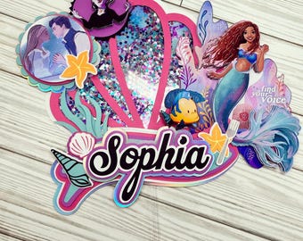 May include: A pink and purple mermaid-themed cake topper with a seashell, a mermaid, a fish, and the name "Sophia" in iridescent letters. The topper also includes a quote: "Find your voice".