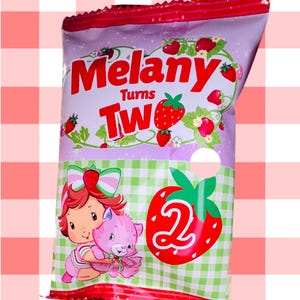 May include: A colorful bag with the words "Melany Turns Two" in red. The bag features a cartoon girl holding a pink cat, strawberries, and a large number "2" inside a strawberry. The background is a gingham pattern.