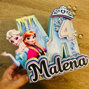 May include: A custom-made birthday decoration featuring the name "Malena" and the number "4". The design incorporates images of Elsa and Anna from the movie Frozen, with a light blue and white color scheme. The decoration includes a crown and snowflake accents.