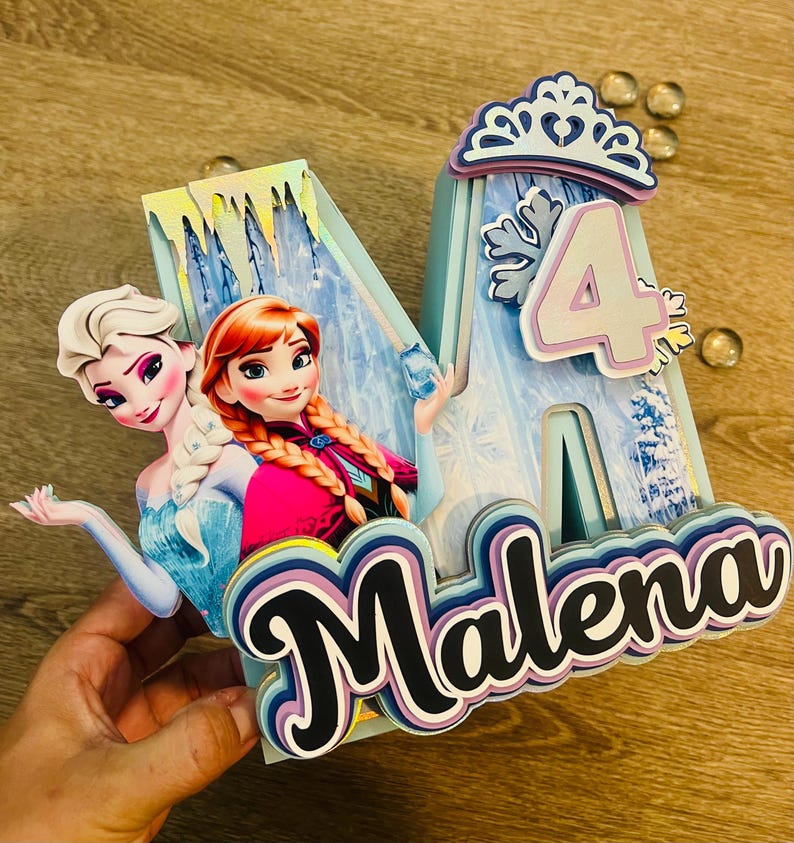 May include: A custom-made birthday decoration featuring the name "Malena" and the number "4". The design incorporates images of Elsa and Anna from the movie Frozen, with a light blue and white color scheme. The decoration includes a crown and snowflake accents.