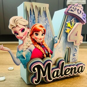May include: A light blue, layered birthday decoration featuring Elsa and Anna from Frozen. The decoration includes the number 4, a crown, and the name "Malena" in a layered, colorful font. The background has a winter theme.