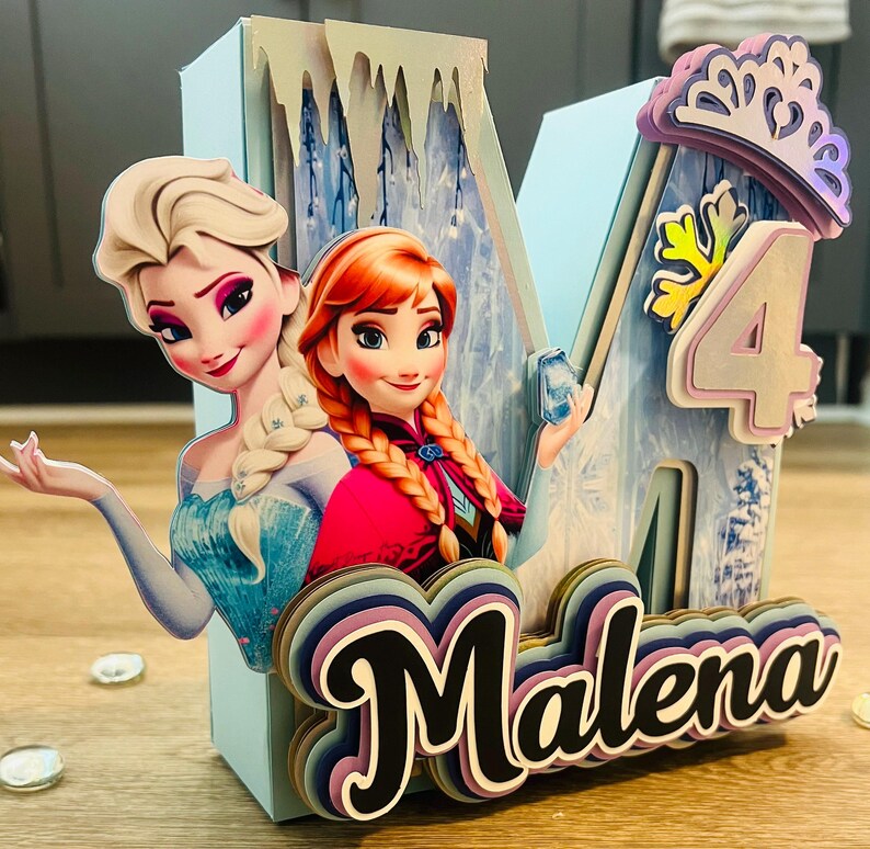 May include: A light blue, layered birthday decoration featuring Elsa and Anna from Frozen. The decoration includes the number 4, a crown, and the name "Malena" in a layered, colorful font. The background has a winter theme.
