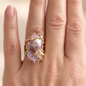 May include: A gold ring featuring a large, irregular, iridescent pink baroque pearl as the centerpiece. The pearl is wrapped in a textured gold wire setting that incorporates several small, round, clear inset stones along the band.